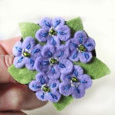 a hand holding a small purple flower brooch with green leaves and beads on it