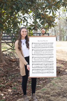 a woman standing in front of a tree holding up a large sheet of musical notation