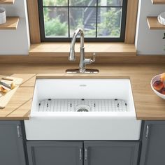 a white kitchen sink sitting under a window next to a bowl of fruit on a counter