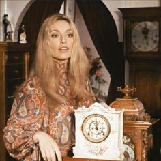 a woman standing in front of a clock with long blonde hair and wearing an ornate dress