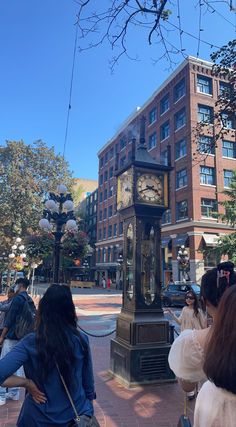 people are standing in front of a clock tower on the side of a street with tall buildings behind them