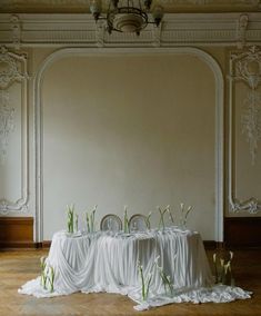 the table is covered with white cloths and green plants in front of an ornate wall