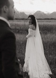 a woman in a wedding dress walking through a field with a man looking at her