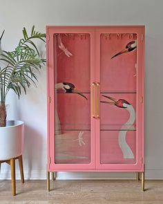 a pink cabinet with birds painted on the glass doors and two plants in a potted planter