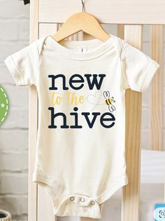 a white baby bodysuit with the words new to the hive on it hanging from a wooden crib