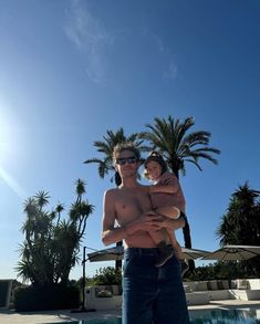 a man holding a child in front of a swimming pool with palm trees behind him