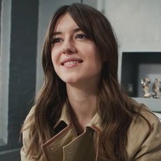 a woman with long hair wearing a trench coat and smiling at the camera while sitting down