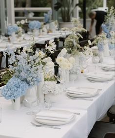 a long table is set with white and blue flowers in vases, plates and napkins