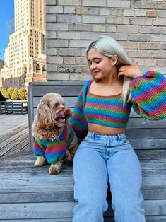 a woman sitting on a wooden bench next to a dog wearing a rainbow striped sweater