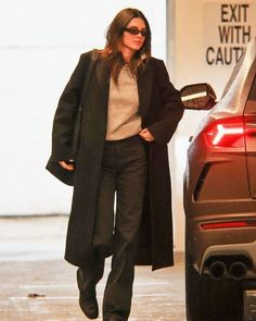 a woman in black coat and pants walking towards a car with her hand on her hip