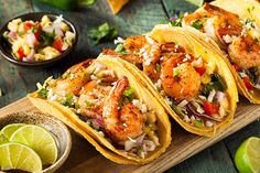 three shrimp tacos on a cutting board with limes, salsa and tortillas