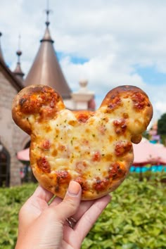 a person holding up a mickey mouse pizza in front of a castle like area with bushes