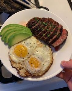 a white plate topped with steak, an egg and sliced avocado on top of it