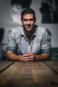 a man sitting at a wooden table with his hands folded in front of the camera