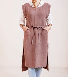 a woman standing in front of a white wall wearing a brown linen apron and jeans