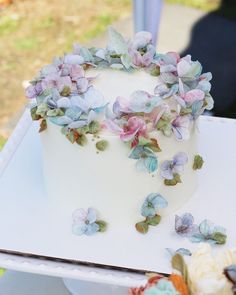 a white cake with purple and blue flowers on it sitting on a table in the grass