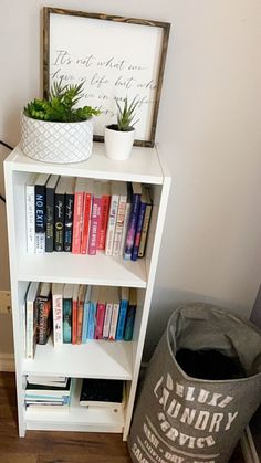 a white book shelf with books and plants on it next to a gray trash can