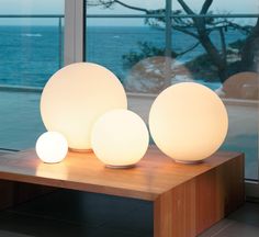 three white lamps sitting on top of a wooden table next to a window with the ocean in the background