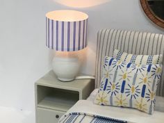 a lamp sitting on top of a night stand next to a white chair with blue and yellow pillows