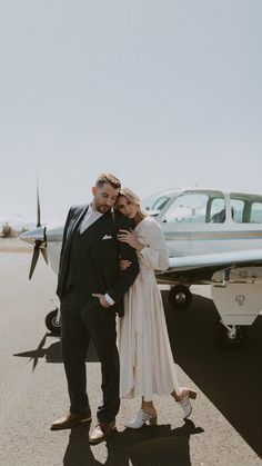 a man and woman standing in front of an airplane on the tarmac with their arms around each other