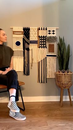 a woman sitting on a chair in front of a wall hanging with weavings and potted plants