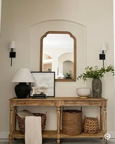 a wooden table topped with baskets under a mirror next to a lamp and vases