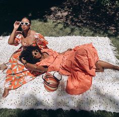 two women laying on a white blanket in the grass, one wearing sunglasses and holding a cell phone to her ear