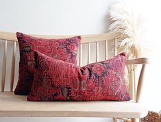 two red pillows sitting on top of a wooden bench next to a white vase with feathers