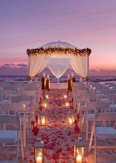 an outdoor wedding setup on the beach with candles lit up and decorated in white linens