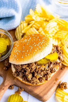 a sloppy joe sandwich with pickles and chips on a cutting board next to it