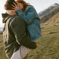 a man and woman embracing each other while standing in a field with mountains behind them