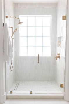 a white tiled shower with gold fixtures in the shower stall and window behind it, along with two towels hanging on hooks