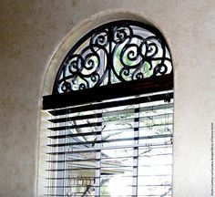 a window with an iron design over the top and blinds on both sides, in front of a white wall