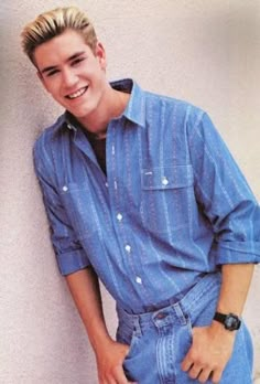 a young man with blonde hair and blue shirt leaning against a wall smiling at the camera