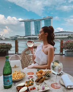 a woman sitting at a table with food and drinks in front of her, overlooking the water