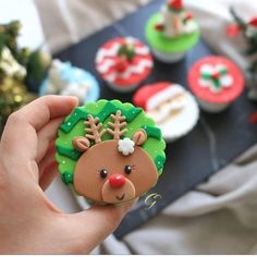 a hand holding a decorated christmas cookie in front of cupcakes on a tray