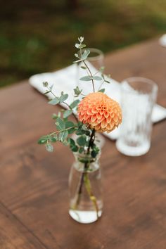 an orange flower sits in a vase on a wooden table with white napkins and glasses