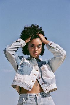 a young woman with curly hair wearing a jean jacket and denim shorts standing in front of a blue sky