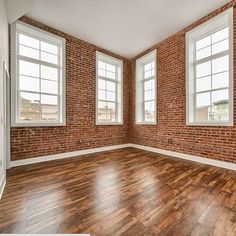 an empty living room with brick walls and hardwood floors is pictured in this image, there are three windows on the far wall