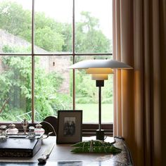 a desk with a lamp and pictures on it in front of a large window that overlooks a lush green yard