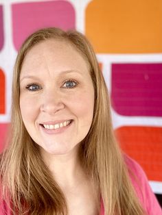 a woman with long blonde hair and blue eyes smiles in front of a colorful wall