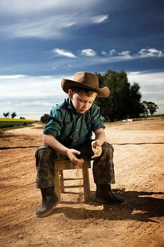 a young boy sitting on top of a wooden chair wearing a cowboy hat and holding something in his hand