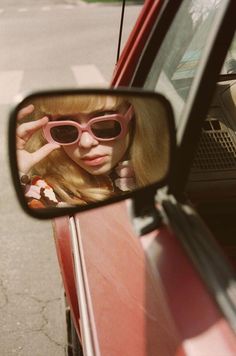 a woman wearing pink sunglasses looking in the side mirror of a red car with her hand on her head