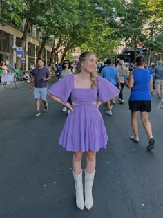 a woman in a purple dress and white boots is standing on the street with her hands on her hips