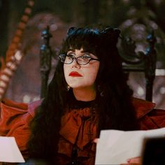 a woman with long black hair and glasses sitting in a chair wearing red clothes, reading papers