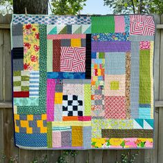 a multicolored quilt hanging on a wooden fence next to a tree and bushes