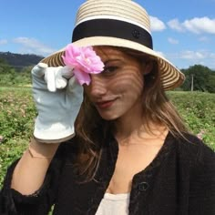 a woman wearing a hat and gloves holding a pink flower in her left hand while looking at the camera