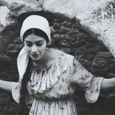 an old photo of a woman in a dress and head scarf standing next to a stone wall