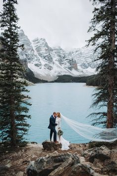 a bride and groom kissing in front of a lake surrounded by snow - capped mountains