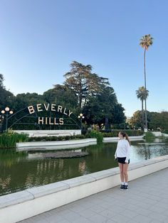 a woman standing in front of beverly hills sign with palm trees and water behind her